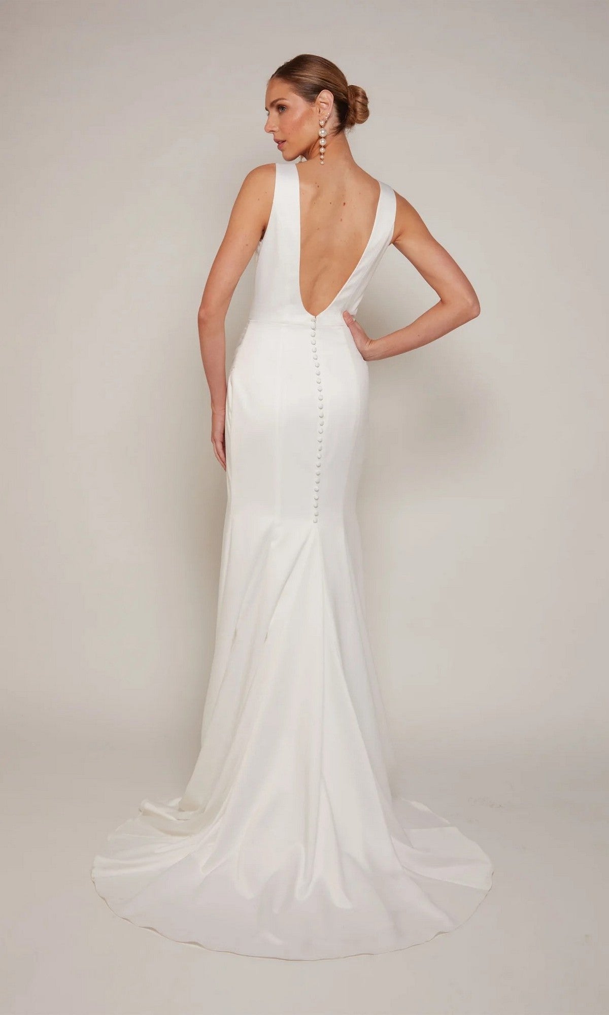 The bride wears the Alyce 7092, a fitted, floor-length white formal dress with a deep V-back, button details, and a slight train, posing against a plain background.